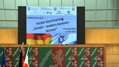 Über dem rednerpult hängt ein Monitor mit dem Titel der Veranstaltung "Israel-Seminartag. Isreal anders kennenlernen"