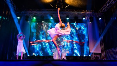 Showauftritt von 3 Tänzerinnen. Mitig springt eine Ballerina in die Höhe, wobie sie ihr linkes Bein hinten hochzieht und gleichzeitig ihren Rücken nachhinten wölbt und den linken Arm nach oben gestreckt hat.