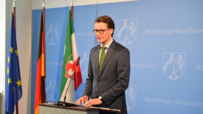 Verkehrsminister Wüst vor blauer NRW-Wand an einem Rednerpult, links neben ihm die Flaggen von Europa, Deutschland und NRW.