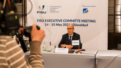 Ein Herr Im Anzug und Krawatte sitz an einem Tisch. Links im Bild sieht man einen Kameramann.