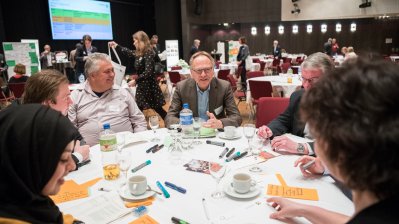 Sieben Menschen sitzen im Gespräch an einen runden Tisch.