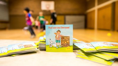 Ein Pixi Buch steht aufgestellt auf einem Tisch. Im Hintergrund sieht man Kinder,die in einer Sporthalle spielen.