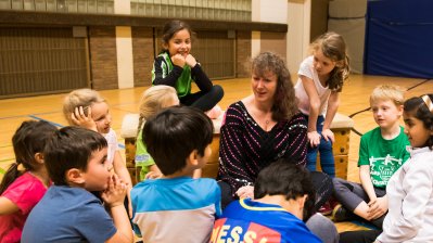 Staatssekretärin Andrea Milz sitzt umringt von Grundschulkindern in einer Sporthalle. IN ihrer Hand hält sie ein Pixi-Buch.