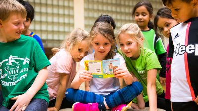 Eine Gruppe vo Grundschulkindern schauen sich ein Pixi-Buch an. Im Hintergrund sieht man die Glaskachelwand der Sporthalle.