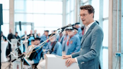 Ministerpräsident Wüst hält eine Rede, im Hintergrund ein Seemann-Chor