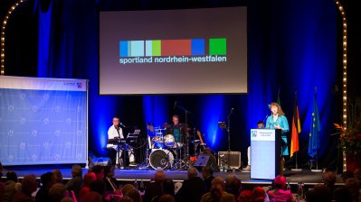 Staatssekretärin Andrea Milz am Rednerpult rechtsseitg auf einer Bühne. Links sitzt eine Musikband. Vor der Bühne sitzt das Publikum.