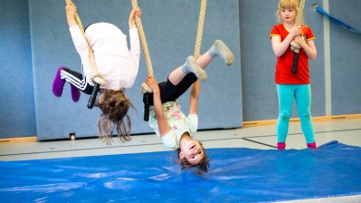 2 Mädchen in weißen T-Shirts und schwarzer Hose hängen kopfüber an den Tauen. 1 Mädchen mit rotem T-shirt und hellblauer Hose steht etwas abseits und hält ihr Tau fest.