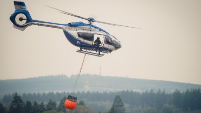 Zu sehen ist ein Löschhubschrauber der Polizei vor einem verrauchten Waldgebiet