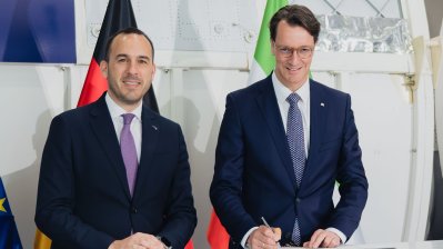 Ministerpräsident Wüst und ein Mann halten NRW-Mappen in die Kamera