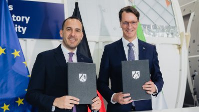 Ministerpräsident Wüst und ein Mann halten das unterzeichnete Papier NRW-Mappen in die Kamera