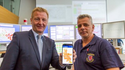 Innenminister Ralf Jäger zeigt auf dem Mobiltelefon neben einem Feuerwehrmann die Notfall-, Informations- und Nachrichten-App NINA 