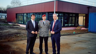 zwei Männer mit Anzug und ein Mann in Bundeswehr-Uniform stehen vor einem roten Gebäude