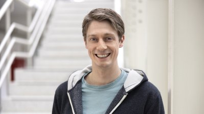 Ein Mann mit hellblauem Tshirt und dunkelblauer Kapuzenjacke steht lächelnd vor einem Treppenaufgang