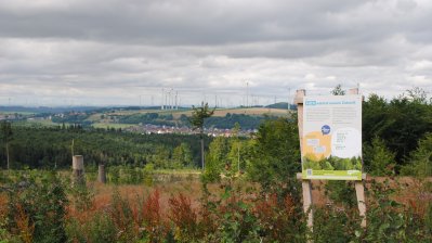 Eine Hinweistafel steht auf einem Hügel an einem Waldrand mit Ausblick in die Ferne