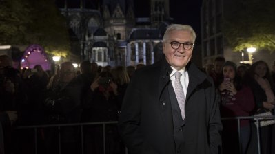 Bundespräsident Steinmeier in Aachen bei Nacht. Im Hintergurnd rechts und links leutchten Straßenlichter, deren Licht die Blätte der danebenstehenden Bäume anleuchten.