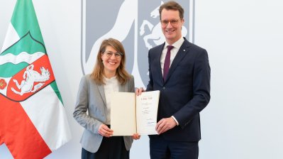 Rechts neben der NRW-Fahne steht Verena Schäffer mit langen blonden Haaren und Brille im grauen Sakko und weißem Hemd lächelnd neben MP Wüst im schwarzen Anzug mit roter Krawatte