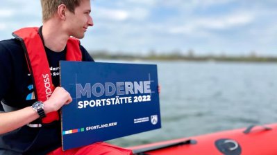 Ein junger Mann sitzt auf einem Schlauchboot und hält ein blaues Schild 