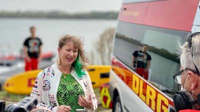 Eine Frau spricht vor einer Kamera, im Hintergrund ist ein Rettungswagen des DLRG