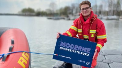 Ein Mann kniet auf einem Steg neben einem Schlauchboot und hält ein blaues Schild 