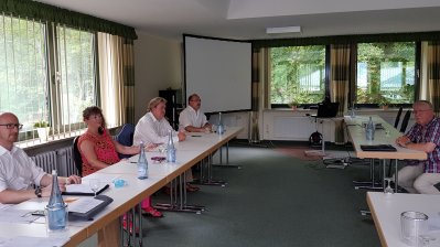 Staatssekretärin Milz sitzt mit drei weiteren Herren an einer Tischreihe in einem Saal, gegenüber sitzt ein Herr im schwarzweiß-kariertem Hemd.