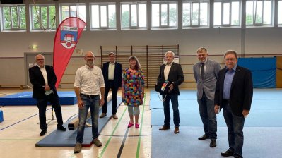 Staatssekretärin Andrea Milz steht mit mehreren Vereinsmitgliedern in einer Turnhalle, überall sidn verstreut Matten auf dem Boden verteilt.