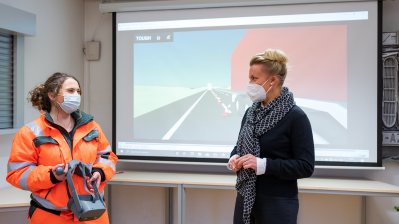 Eine Straßenwärterin im orangenen Overall zeigt der Ministerin eine VR-Brille vor einer Leinwand