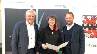 Staatssekretärin Milz steht zwischen zwei Herren und übergibt den Zuwendungsbescheid.