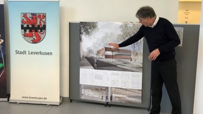 Rechts steht ein Herr im weißen Hemd, schwarzen Pullover und schwarzer Hose auf ein Bild zeigend, dabei hält er einen Vortrag. Links sieht man das Wappen von der Stadt Leverkusen mit rotem Löwen auf weißen Grund und einem schwarzen Balkenmuster mittig quer verlaufend.
