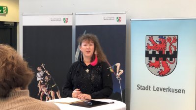 Staatssekretärin Andrea Milz sitzt an einem Tisch, vor ihr sitzt ein Publikum. Rechts sieht man das Wappen von Leverkusen, den roten Löwen auf weißen Grund und einem schwarzen Balkenmuster mittig quer verlaufend.