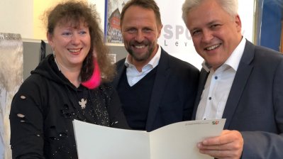 Staatssekretärin Milz steht links neben zwei Herren von der Stadt Leverkusen, gemeinsam halten sie den Zuwendungsbescheid.