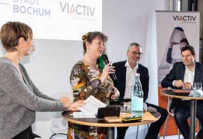 Staatssekretärin Andrea Milz mit einem Mikrofon in der Hand sitzt mit Teilnehmern der Pressekonferenz auf Hockern an Rundtischen. Hinter ihnen eine weiße Rückwand mit einer Präsentation.