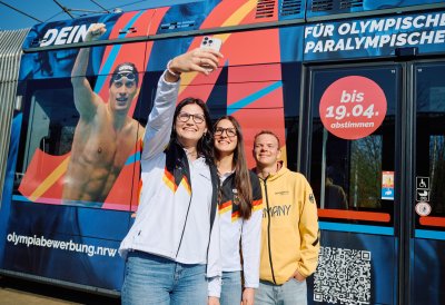 3 Sportlerinnen und Sportler machen ein Selfie vor der Straßenbahn