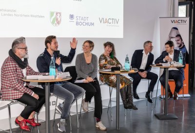 Staatssekretärin Andrea Milz sitzt mit Teilnehmern der Pressekonferenz auf Hockern an Rundtischen. Hinter ihnen eine weiße Rückwand mit einer Präsentation.