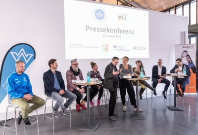 Staatssekretärin Andrea Milz sitzt mit Teilnehmern der Pressekonferenz auf Hockern an Rundtischen. Hinter ihnen eine weiße Rückwand mit einer Präsentation.
