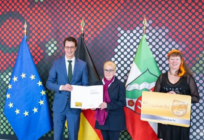 Ein Mann und zwei Frauen stehen vor der Europa-, Deutschland- und NRW-Flagge