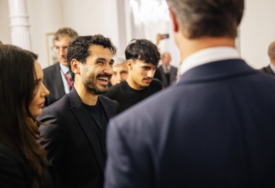 Fußballer İlkay Gündoğan trägt einen dunklen Anzug und lacht zwischen anderen Personen in Business-Kleidung.