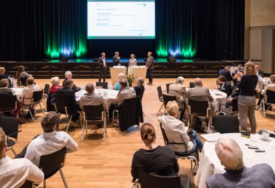 Ein Saal mit vielen, runden, weißen Tischen mit Gästen, weiter vorne ein Bühne  mit einer großen Leinwand.