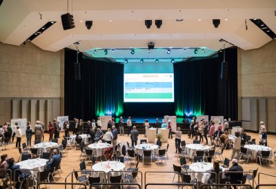 Ein Saal mit vielen, runden, weißen Tischen mit Gästen, weiter vorne ein Bühne  mit einer großen Leinwand.