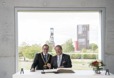 Ministerpräsident und der Oberbürgermeister stehen nebeneinander vor dem Stahlbuch und schauen in die Kamera. Im Hintergrund ist die Zeche Zollverein zu sehen.