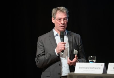 Ein Redner im grauen Anzug und weißem Hemd mit Mikrofon in der Hand am Rednerpult.