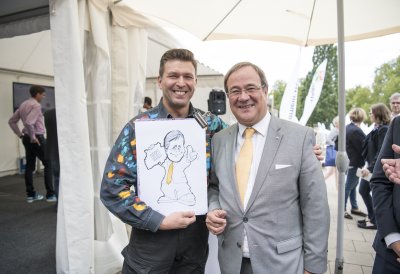 Ein Karikaturist steht mit seiner Zeichnung neben Ministerpräsident Armin Laschet