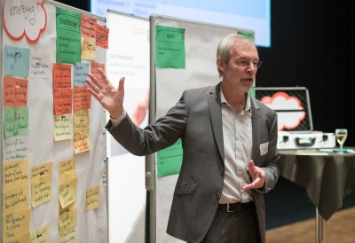 Ein Moderator im grauem Anzug und wießem Hemd zeigt auf eine Pinnwand.