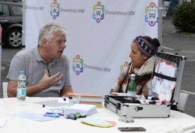 Ein älterer Herr mit einer jungen Dame im Gespräch an einem Tisch sitzend. Im HIntergrund eine Stellwand mit Ehrenamtstour-Logos und dem Ehrenamtstourbus.