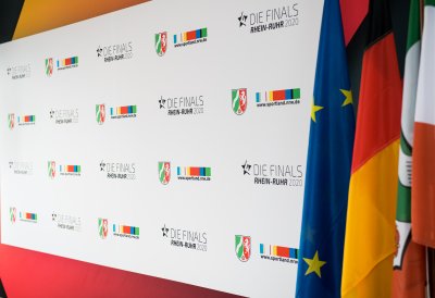 Weiße Plakatwand mit NRW Wappen und Logos von Sportland.NRW, rechts davon hängen die Flaggen von der EU, Deutschalnd und NRW.
