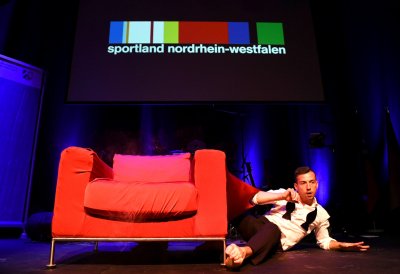 Showakt - Auf der Bühne steht ein großer roter Sessel, rechts daneben liegt ein Mann auf einem Arm abgestüt, den anderen Arm angewinkelt. Er hat ein weißes Oberteil und eine schwarze Hose an.