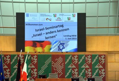 Über dem rednerpult hängt ein Monitor mit dem Titel der Veranstaltung "Israel-Seminartag. Isreal anders kennenlernen"