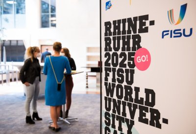 Drei Damen stehen draußen an einem runden Stehtisch. Rechts sieht man von der Rhine Ruhr 2025 FISU,.schwarz auf weiß.