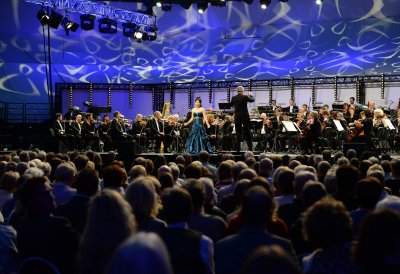 Eine Opernsängerin steht neben dem Dirigenten, im Hintergrund das Orchester, im Vordergrund Köpfe des Publikums