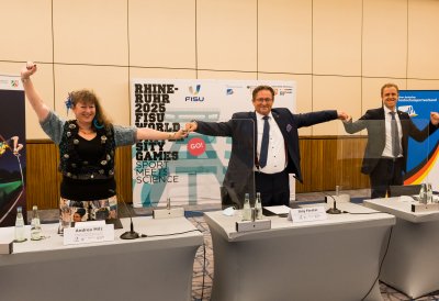 Staatssekretärin Milzsteht mit zwei Herren hinter einer Tischreihe und bilden mit gestreckten Armen eine Kette des Zusammenhalts.