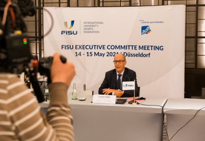 Ein Herr Im Anzug und Krawatte sitz an einem Tisch. Links im Bild sieht man einen Kameramann.